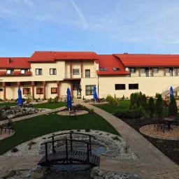 Rozległy budynek hotelowy z czerwonym dachem, otoczony zielonym trawnikiem, ścieżkami z kostki, dekoracyjnymi krzewami i małymi drzewami, niebieskie parasole ogrodowe przy stolikach, mały mostek...