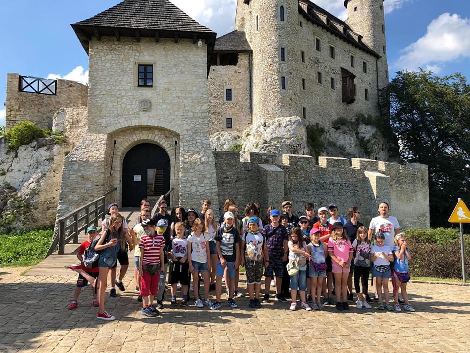 Grupa dzieci i dorosłych na tle średniowiecznego zamku w słoneczny dzień, część osób w maseczkach, wycieczka szkolna.