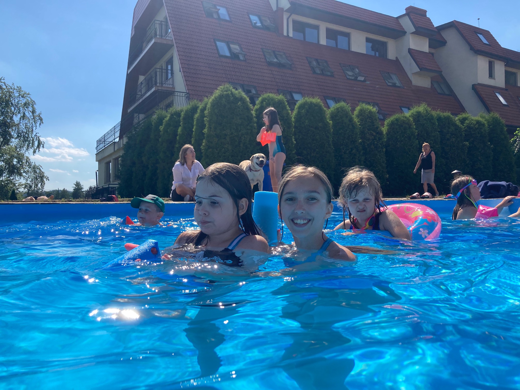 Grupa dzieci w basenie na tle budynku z czerwonym dachem i zielonymi tujami, jedno dziecko z psem i opiekunem stoi obok basenu.