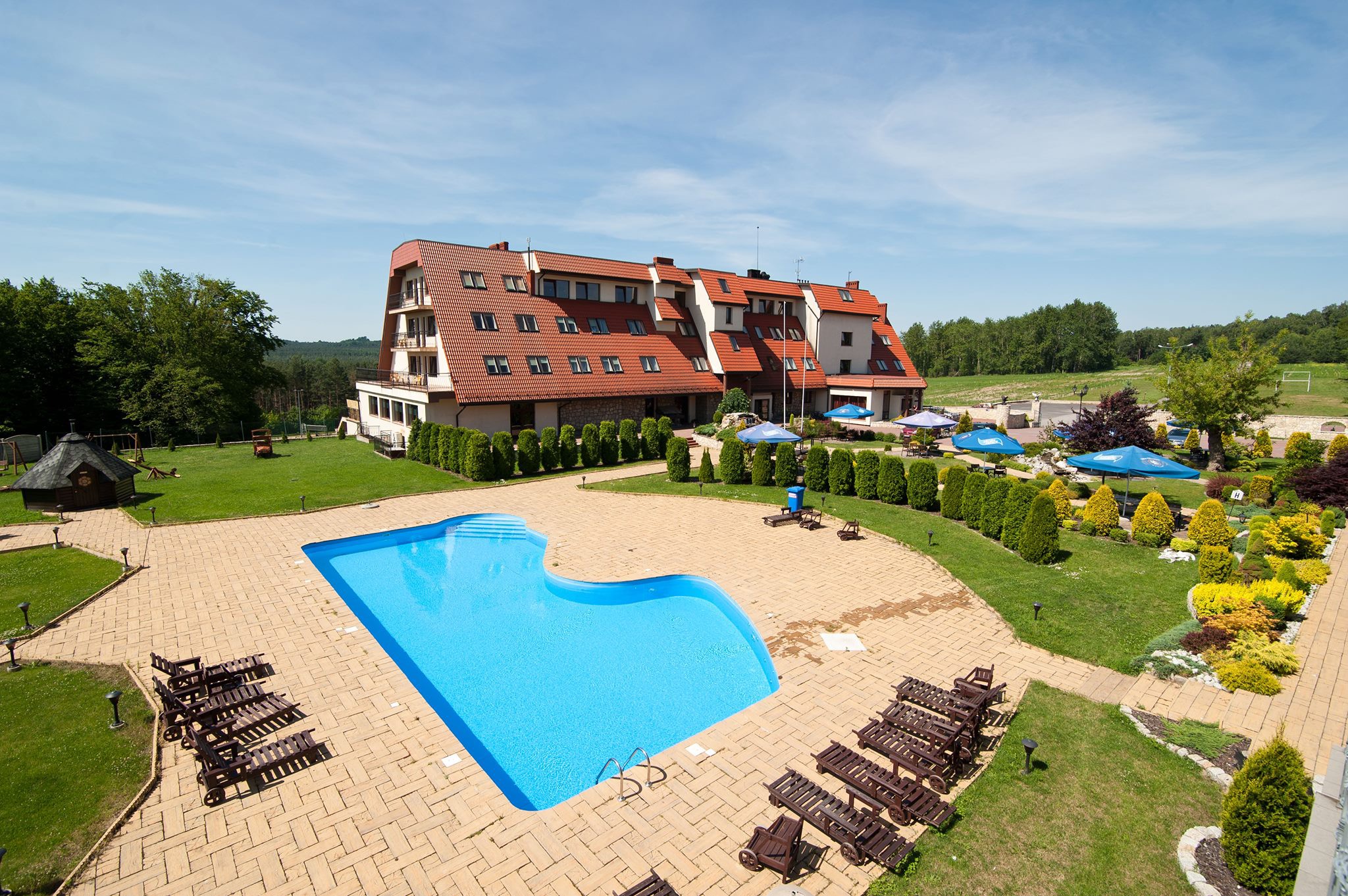 Widok z góry na hotel z basenem o nieregularnym kształcie, otoczony zielenią, leżakami i parasolami.