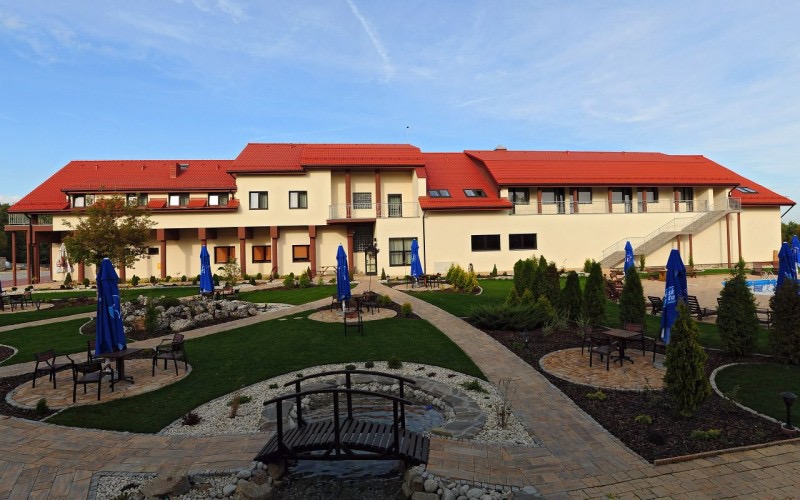 Rozległy budynek hotelowy z czerwonym dachem, otoczony zielonym trawnikiem, ścieżkami z kostki, dekoracyjnymi krzewami i małymi drzewami, niebieskie parasole ogrodowe przy stolikach, mały mostek...