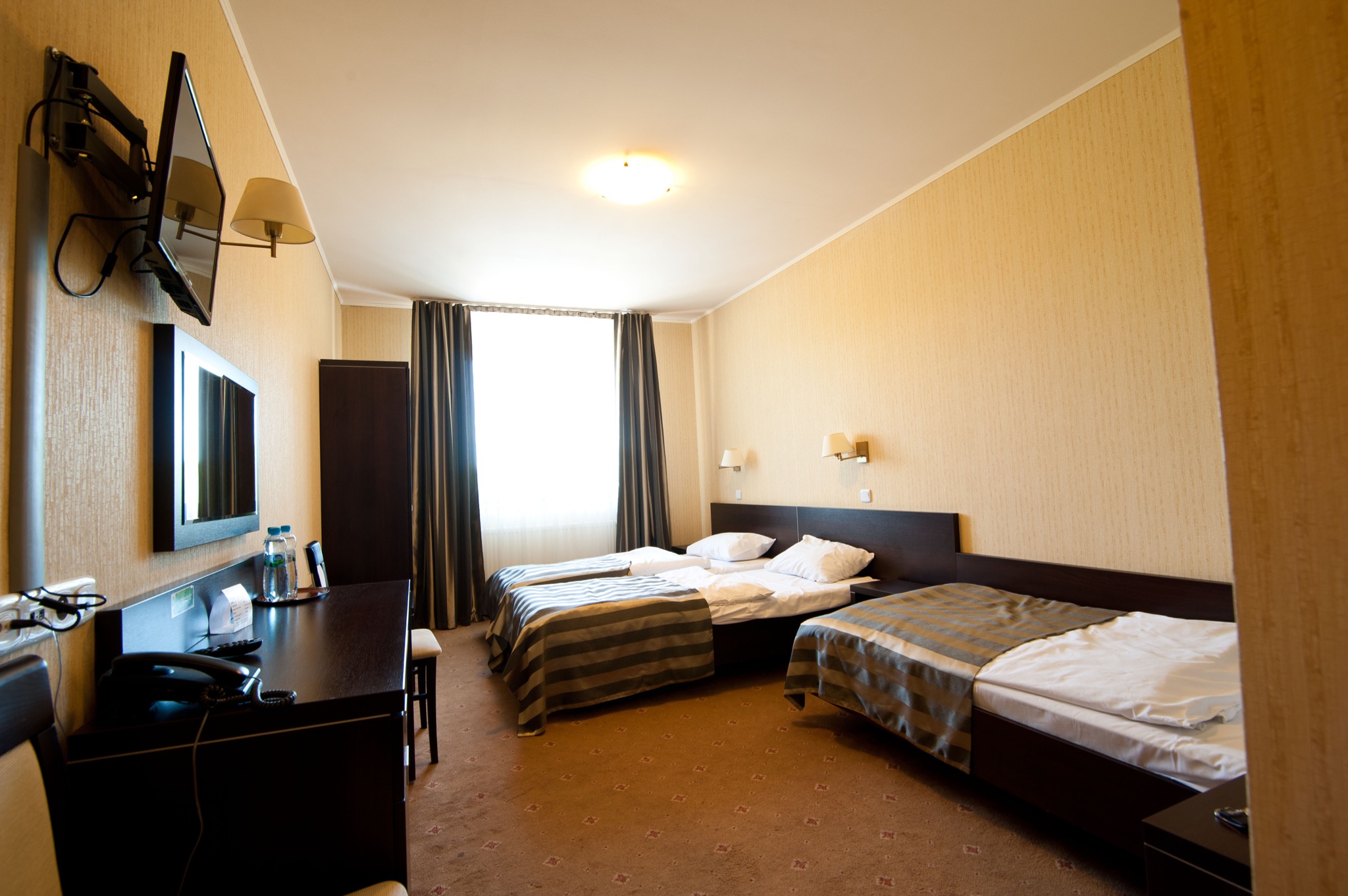Jasny pokój hotelowy z trzema pojedynczymi łóżkami, biurkiem, telewizorem i oknem z ciemnymi zasłonami.