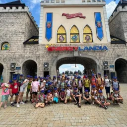 Grupa dzieci w niebieskich czapkach pozuje na tle wejścia do Energylandii, widoczne kamienne mury stylizowane na zamek i logo parku rozrywki.