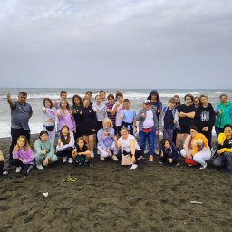 BIURO PODRÓŻY B&B TRAVEL - Grupa dzieci i dorosłych na czarnej, piaszczystej plaży z falami morskimi w tle, wszyscy pokazują kciuki w górę.