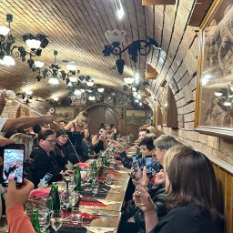 BIURO PODRÓŻY B&B TRAVEL - Wnętrze restauracji z ceglanym sklepieniem, długi stół z nakryciami i siedzącymi gośćmi, część osób robi zdjęcia telefonami, kelnerka nalewa napój.