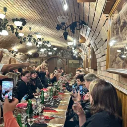 Wnętrze restauracji z ceglanym sklepieniem, długi stół z nakryciami i siedzącymi gośćmi, część osób robi zdjęcia telefonami, kelnerka nalewa napój.