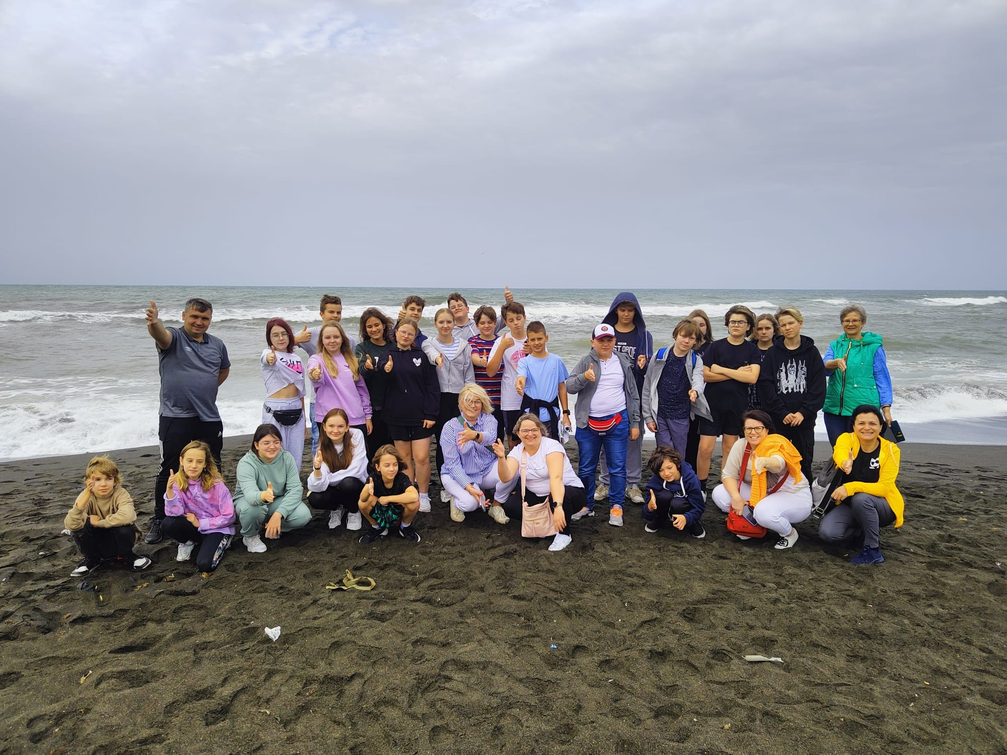 Grupa dzieci i dorosłych na czarnej, piaszczystej plaży z falami morskimi w tle, wszyscy pokazują kciuki w górę.