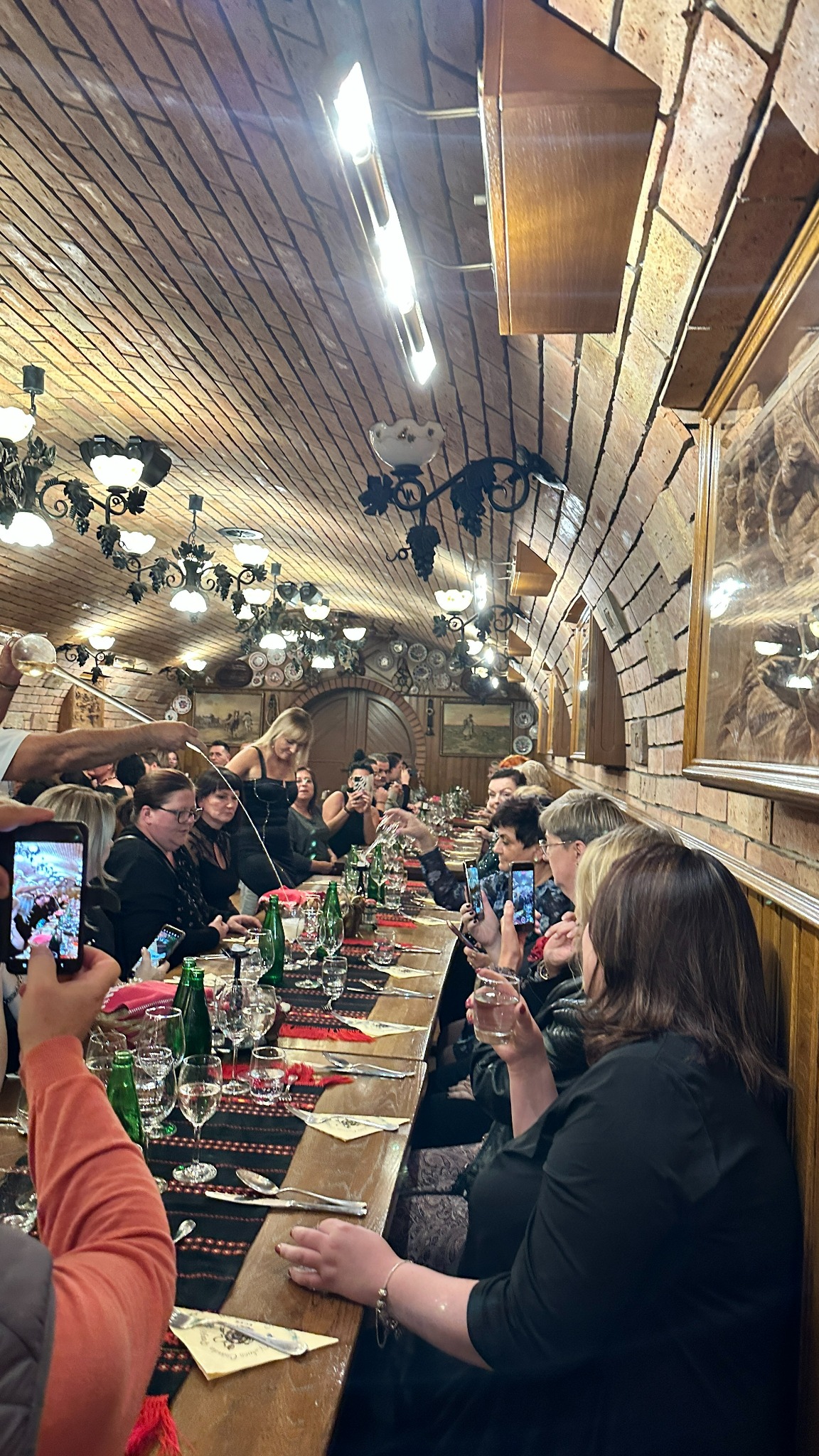 Wnętrze restauracji z ceglanym sklepieniem, długi stół z nakryciami i siedzącymi gośćmi, część osób robi zdjęcia telefonami, kelnerka nalewa napój.