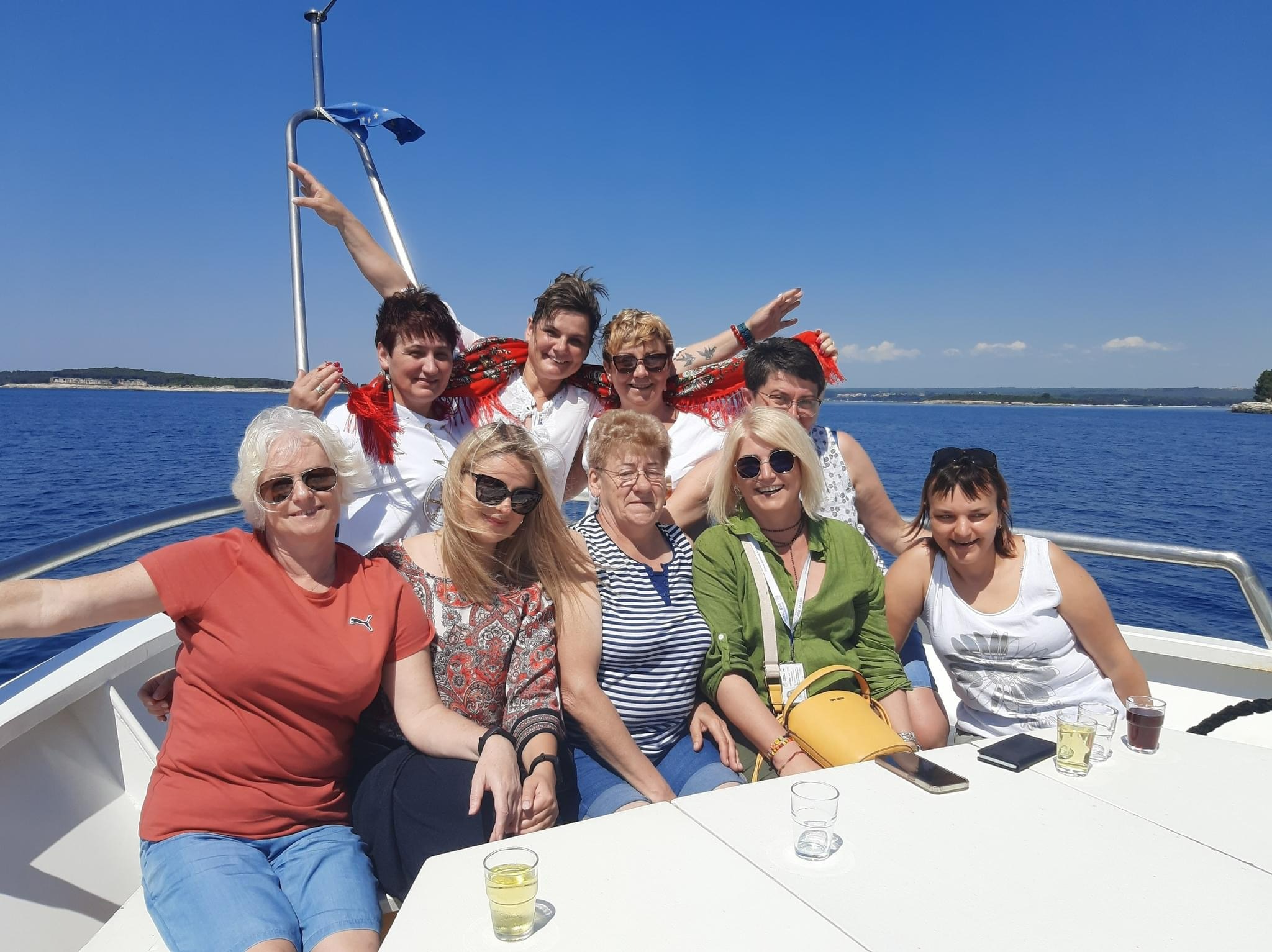 Grupa kobiet w różnym wieku pozuje na pokładzie łodzi podczas rejsu po morzu w słoneczny dzień, widoczne napoje na stole.