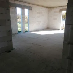 Surowy stan wnętrza domu w trakcie budowy, widoczne ściany z białych bloczków, betonowy strop i wylewka podłogowa, z oknami bez ram wychodzącymi na zewnątrz, widok z wnętrza przez otwór drzwiowy.