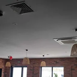 Sufit w lokalu komercyjnym z zamontowaną kasetą klimatyzacyjną, oświetleniem szynowym i dekoracyjnymi lampami wiszącymi z abażurami z rattanu.