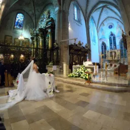 Panna młoda w białej sukni klęczy przed ołtarzem w kościele, ksiądz w tle, bogato zdobione wnętrze z witrażami i kolumnami.