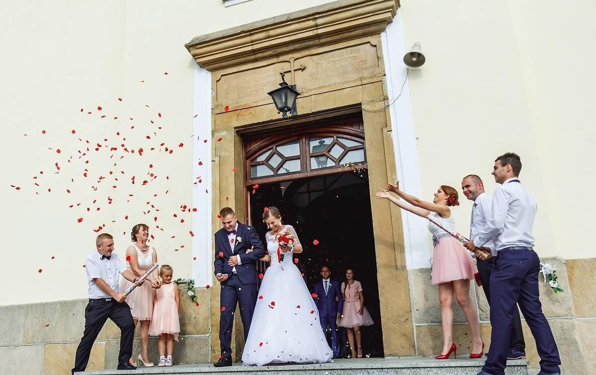 Para młoda wychodzi z kościoła, obsypywana płatkami róż przez gości weselnych; w tle kamienna fasada budynku z inskrypcją.