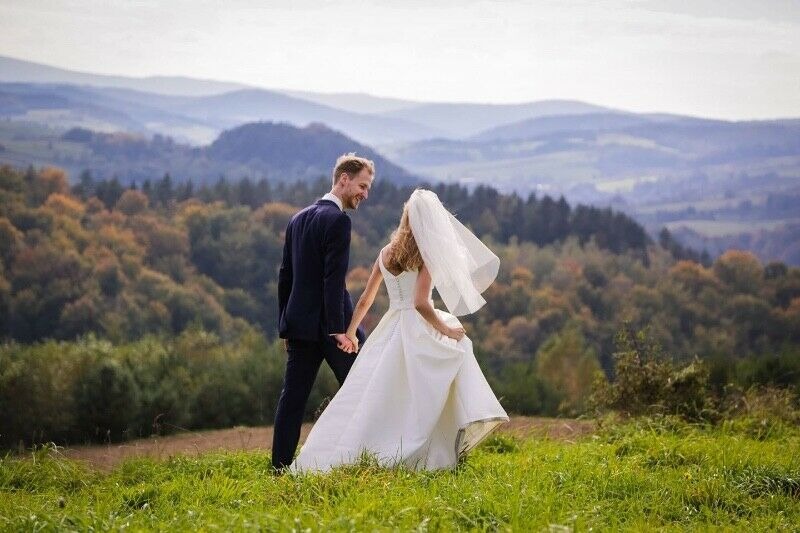 Para młoda, on w granatowym garniturze, ona w białej sukni ślubnej z welonem, idąca po zielonej łące na tle zalesionych wzgórz i gór w oddali.