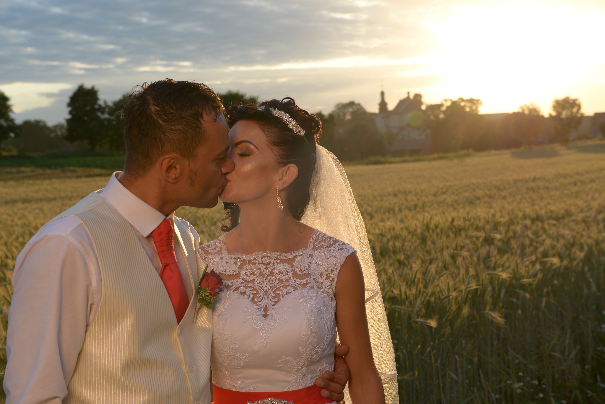 Para młoda całuje się na tle pola zboża o zachodzie słońca, zarys zamku w oddali.