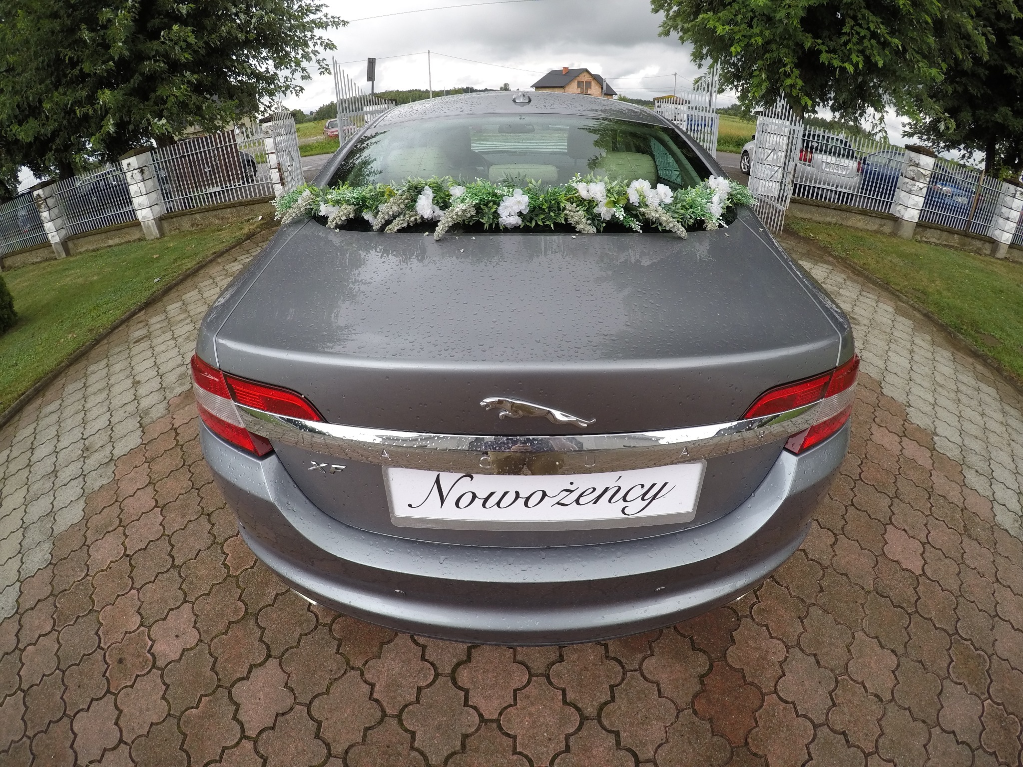 Tył szarego samochodu Jaguar XF z dekoracją kwiatową i tablicą 'Nowożeńcy' na tle kostki brukowej i ogrodzenia.