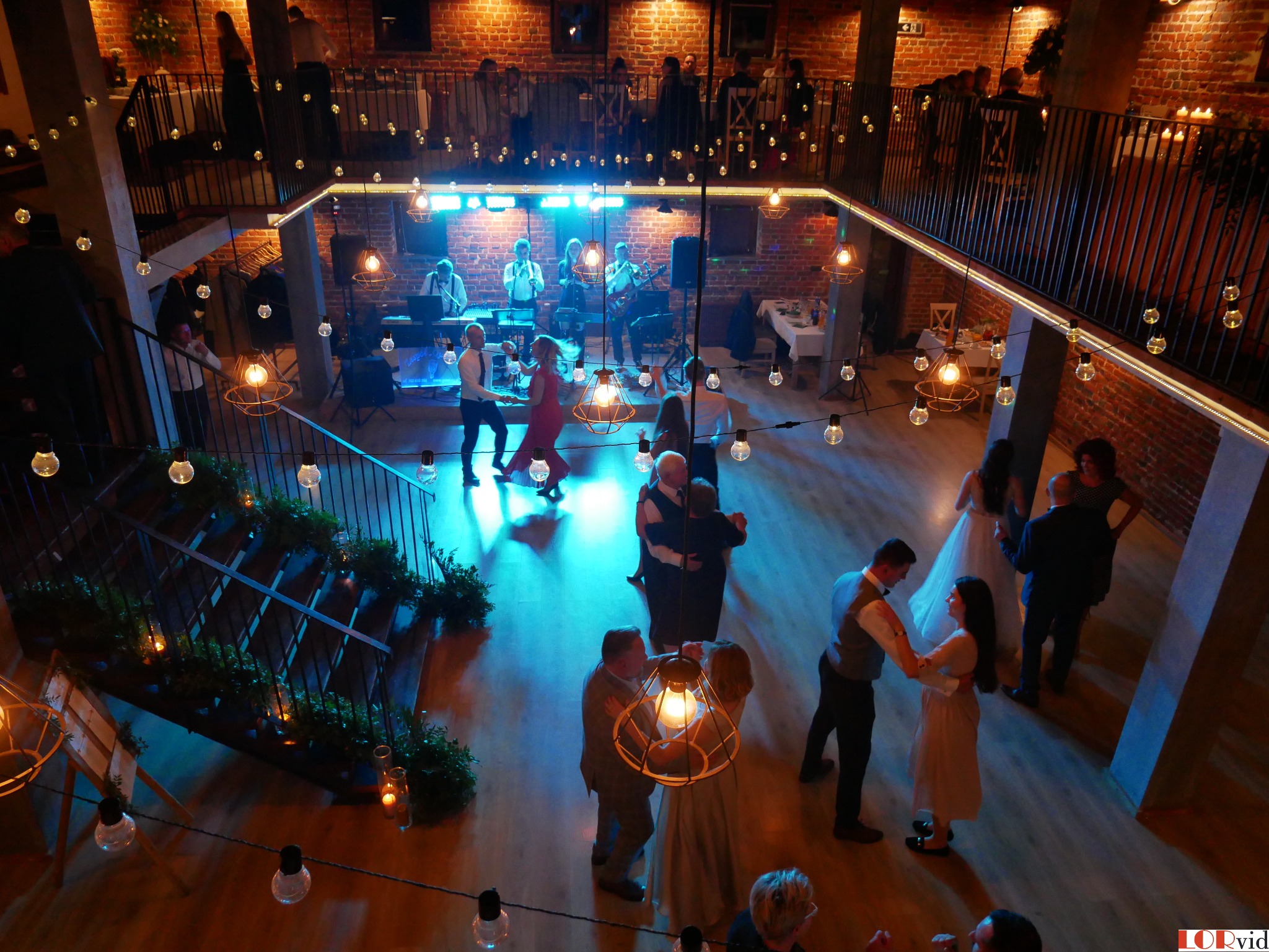 Sala weselna z ceglanymi ścianami, oświetlona girlandami żarówek, z parkietem, na którym tańczą pary, oraz zespołem muzycznym na scenie w tle.