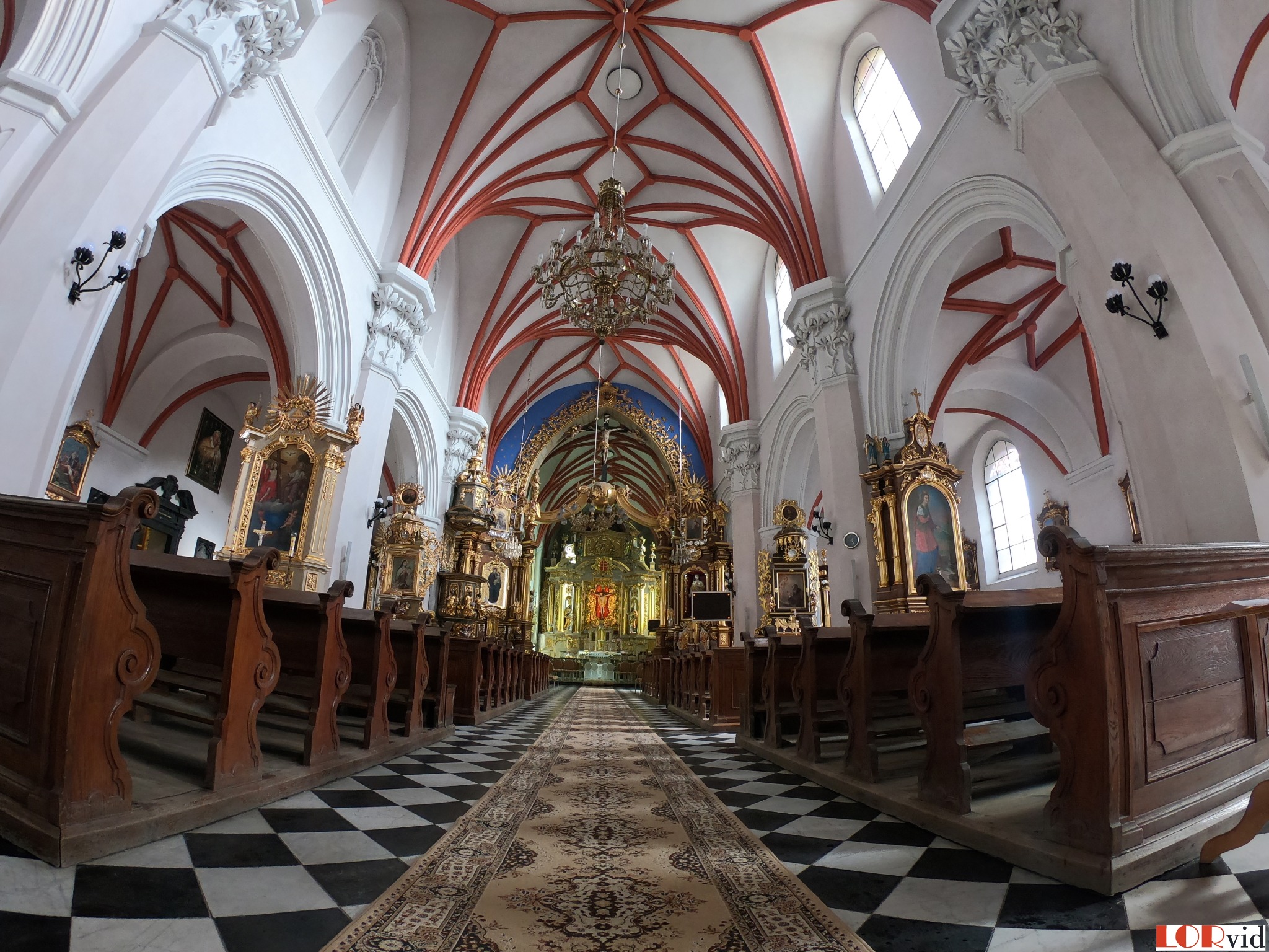 Wnętrze kościoła z perspektywy podłogi, ukazujące nawę główną z ławkami, czarno-białą posadzką, dywanem prowadzącym do ołtarza, bogato zdobiony ołtarz, sklepienie krzyżowe z żyrandolem i boczne...