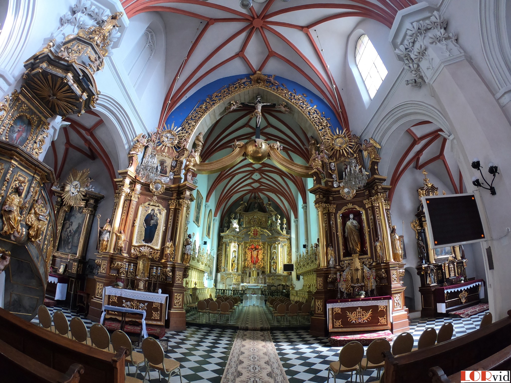 Wnętrze barokowego kościoła z bogato zdobionym ołtarzem, widok z perspektywy rybiego oka, ukazujący sklepienie z czerwonymi żebrami i czarno-białą posadzkę.