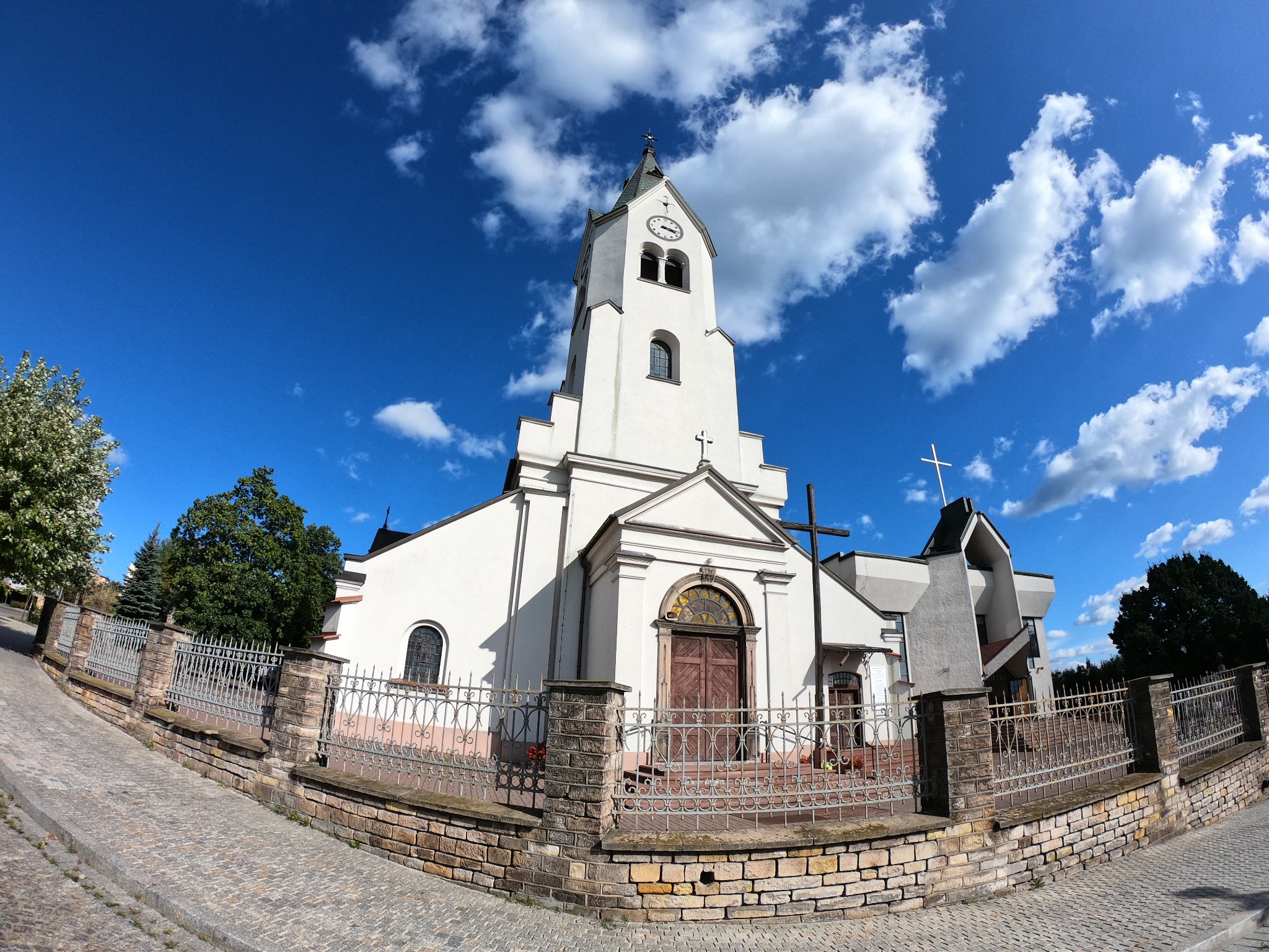 Szerokokątny widok na biały kościół z wieżą zegarową, ogrodzony niskim murem z metalową balustradą, na tle błękitnego nieba z chmurami.