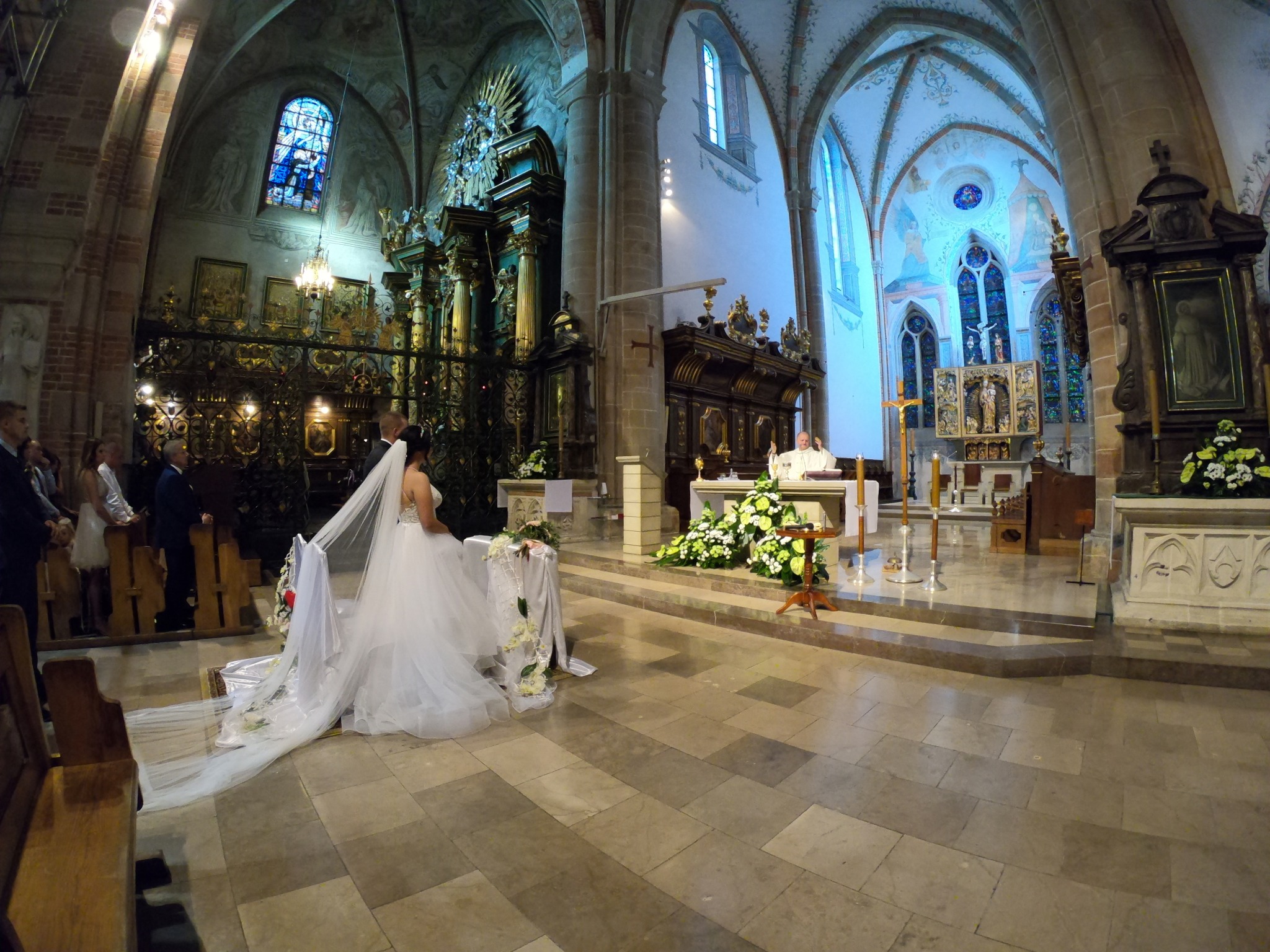 Panna młoda w białej sukni klęczy przed ołtarzem w kościele, ksiądz w tle, bogato zdobione wnętrze z witrażami i kolumnami.