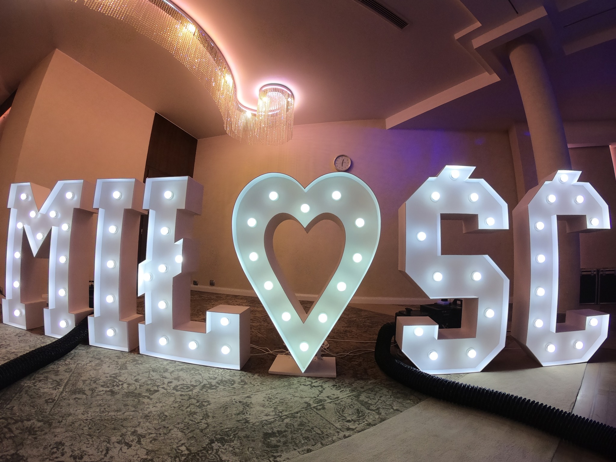 Podświetlane, białe litery układające się w słowo 'MIŁOŚĆ' z sercem zamiast litery 'O', ustawione na podłodze sali bankietowej w Krakowie, z widocznym kryształowym żyrandolem w tle.
