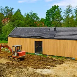 Nowoczesny dom z panelami słonecznymi na dachu w trakcie budowy, z widoczną mini koparką i przygotowanym terenem.