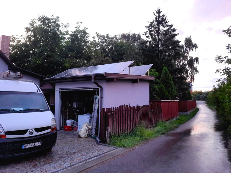 Panele słoneczne na dachu garażu, widok z ulicy po deszczu, z samochodem dostawczym firmy obok.
