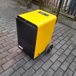 Żółto-czarny przemysłowy osuszacz powietrza z wyświetlaczem i uchwytem transportowym, stojący na kostce brukowej.