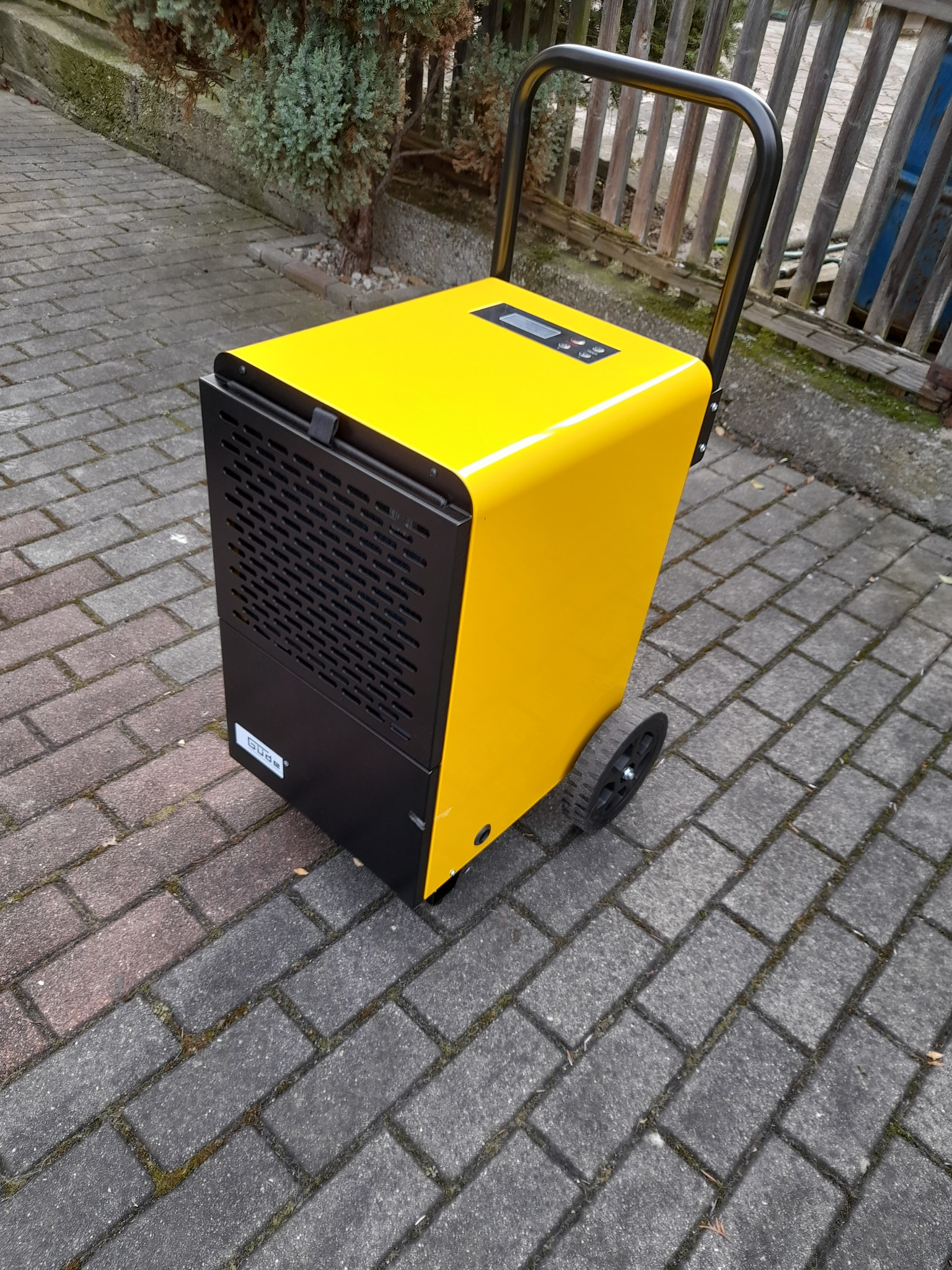 Żółto-czarny przemysłowy osuszacz powietrza z wyświetlaczem i uchwytem transportowym, stojący na kostce brukowej.