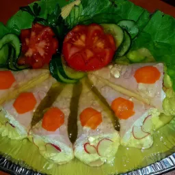 Galaretka z szynką, rzodkiewką, marchewką i kukurydzą w galarecie, udekorowana pomidorami, ogórkami i sałatą na srebrnej tacy.