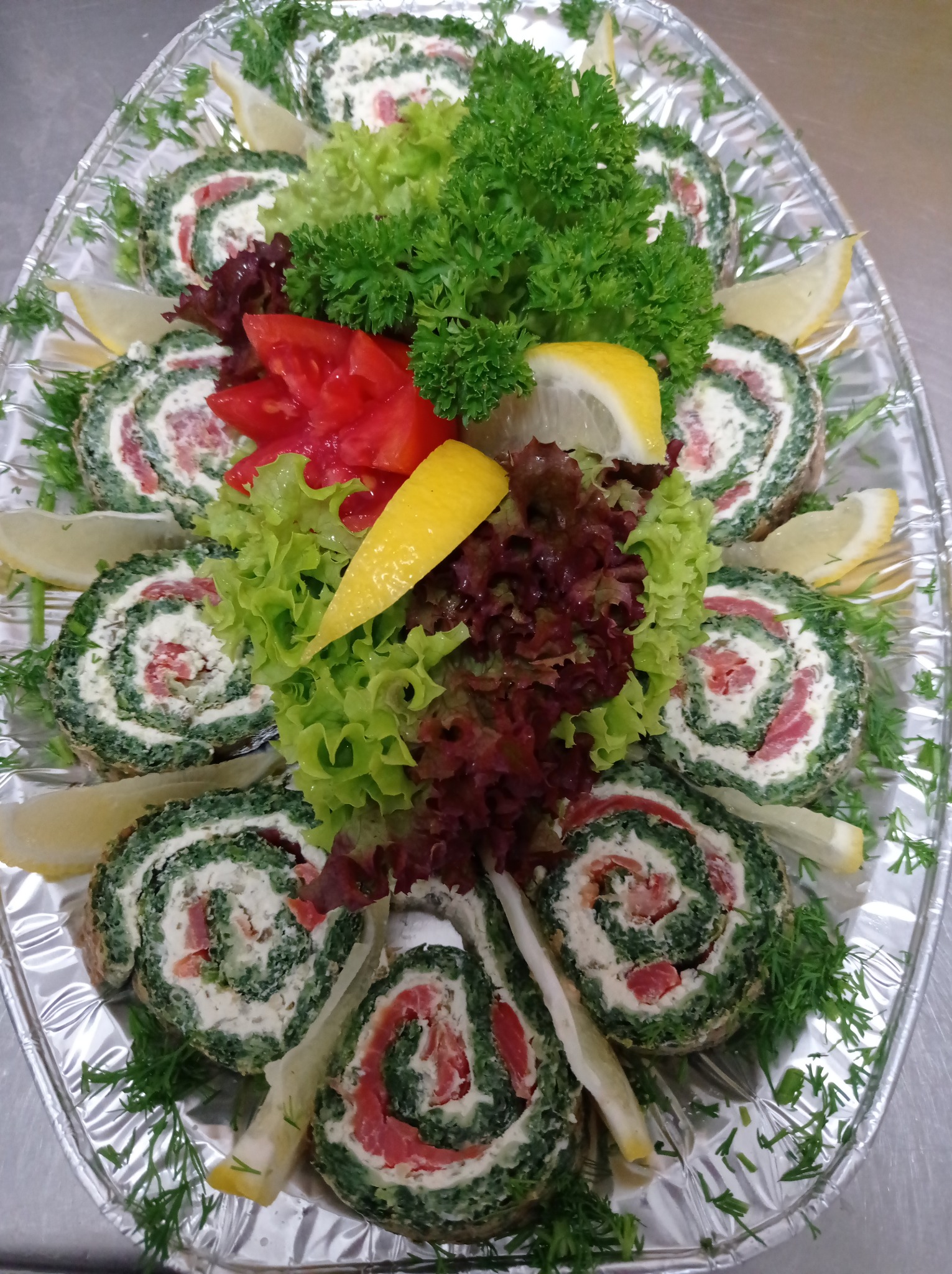 Dekoracyjnie ułożone roladki ze szpinakiem i łososiem na srebrnej tacy, ozdobione cytryną, pomidorem, sałatą i koperkiem.