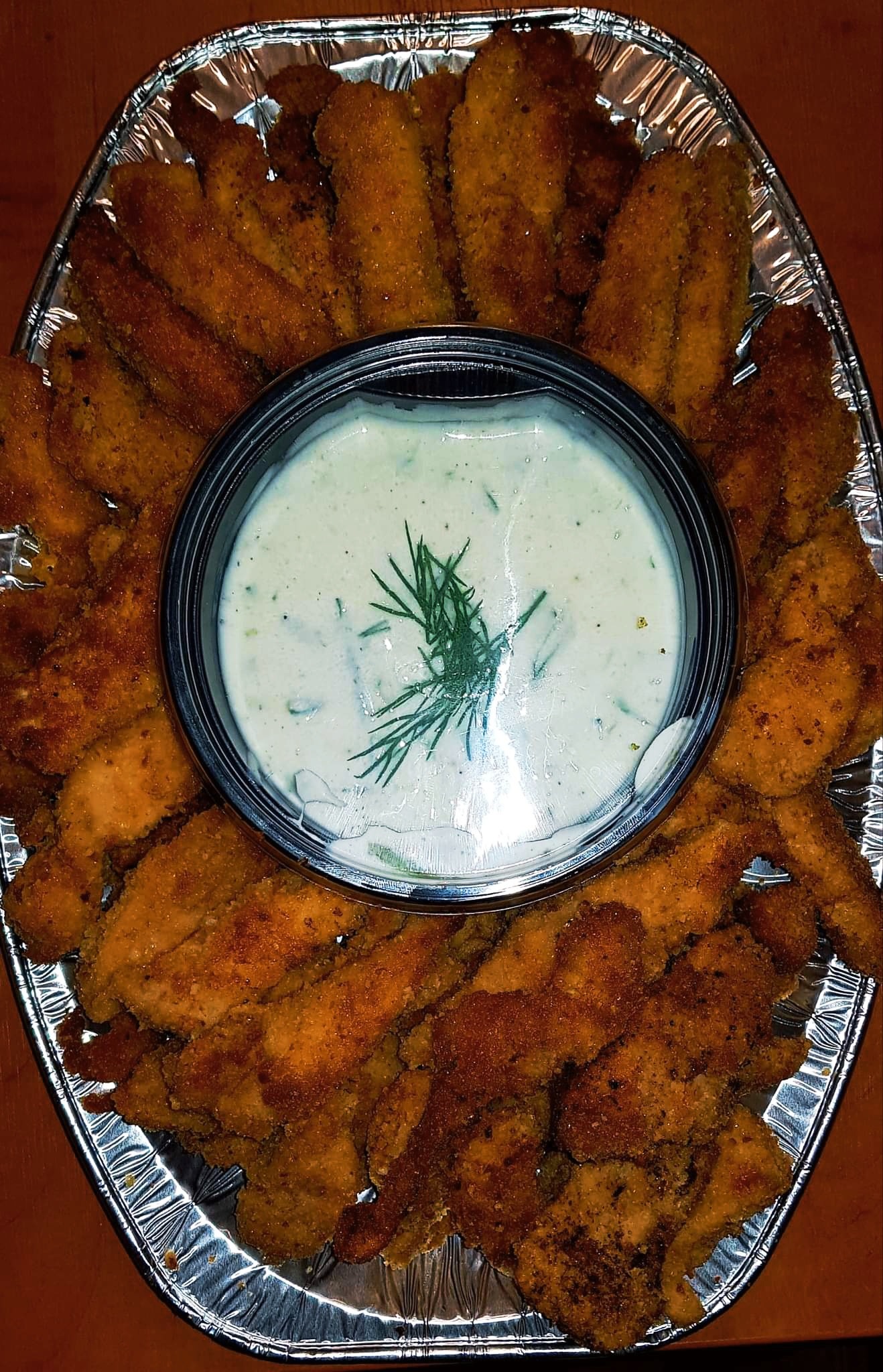 Owalna aluminiowa taca z panierowanymi kawałkami kurczaka ułożonymi wokół okrągłego pojemnika z sosem tzatziki, udekorowanym gałązką koperku.