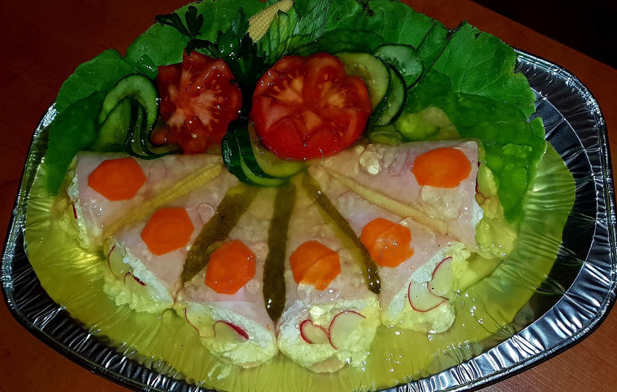 Galaretka z szynką, rzodkiewką, marchewką i kukurydzą w galarecie, udekorowana pomidorami, ogórkami i sałatą na srebrnej tacy.