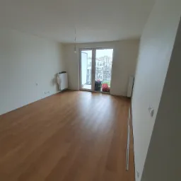 Puste, jasne mieszkanie z panelami podłogowymi, białymi ścianami i balkonem z widokiem na miasto; widoczne punkty elektryczne, grzejnik i wisząca żarówka bez klosza.