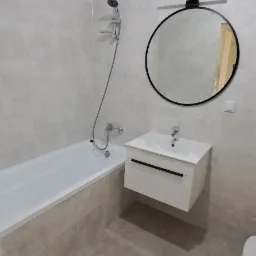 Nowoczesna łazienka w odcieniach szarości z wiszącą białą szafką pod umywalką, okrągłym lustrem z czarną ramą i baterią umywalkową oraz wanną z zamontowaną baterią i słuchawką prysznicową.