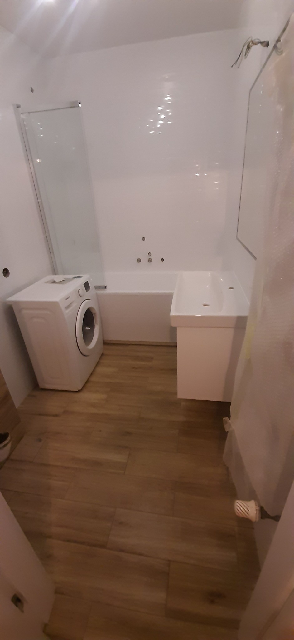 Jasna łazienka z drewnopodobną podłogą, białą wanną z hydromasażem, umywalką na szafce i pralką, widoczne elementy instalacji elektrycznej na ścianie.