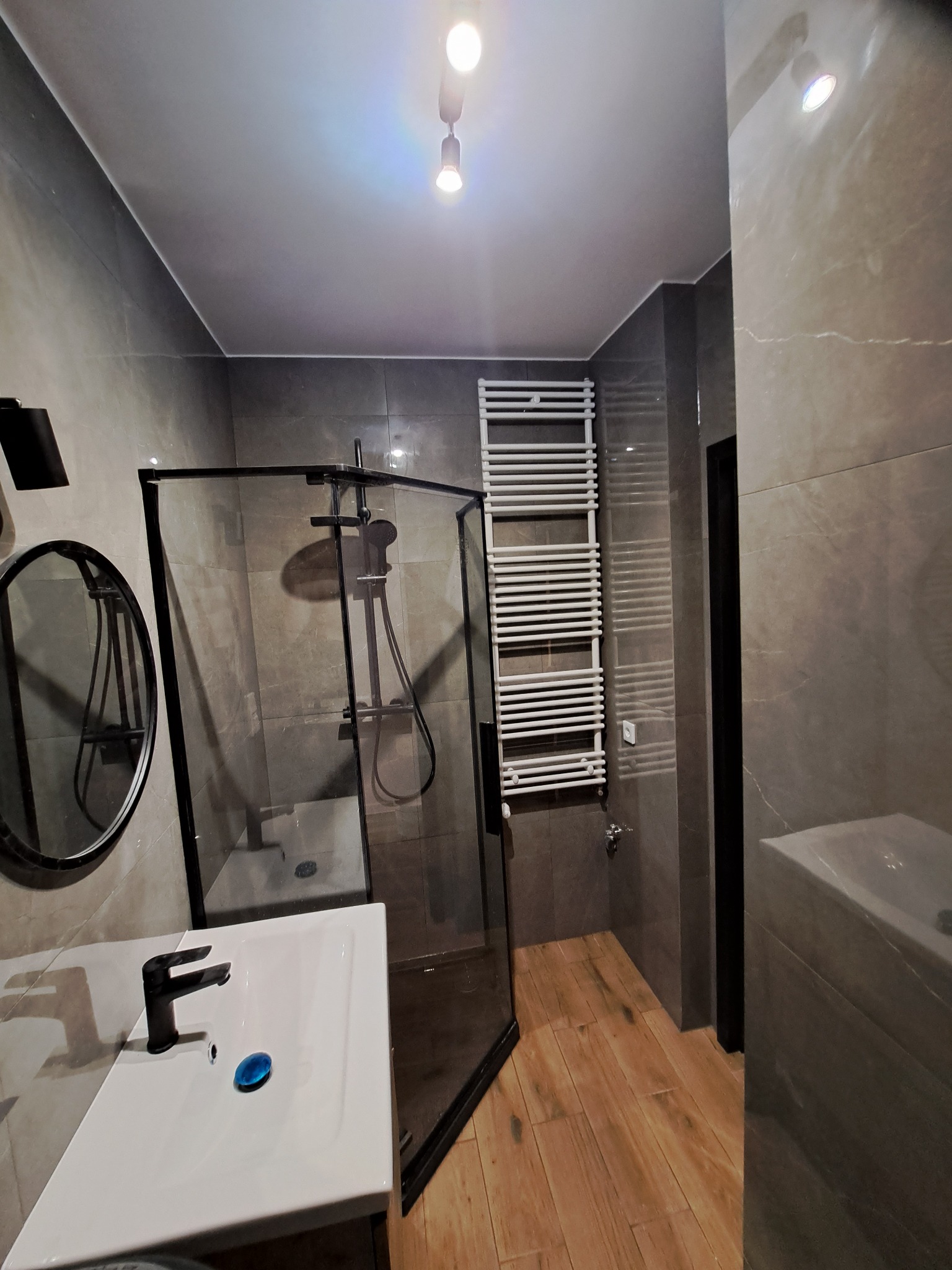 Nowoczesna łazienka z kabiną prysznicową typu walk-in, umywalką z czarną armaturą, okrągłym lustrem i grzejnikiem drabinkowym na tle szarych płytek imitujących marmur i drewnopodobnej podłogi.