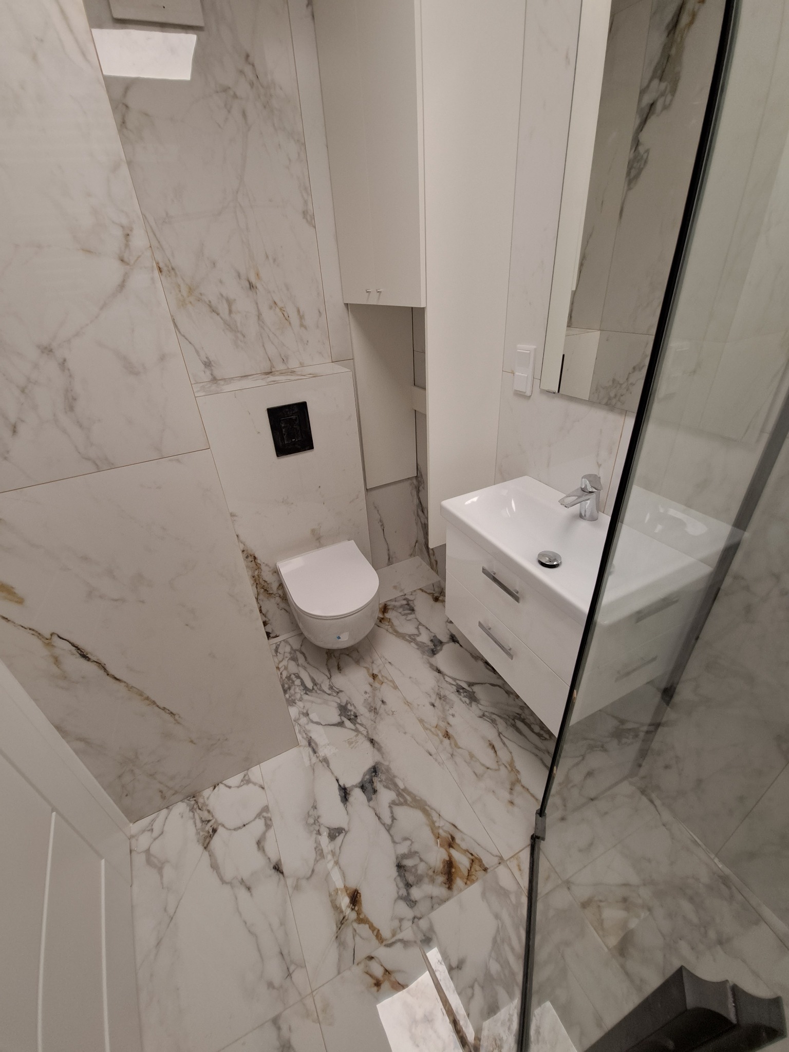 Nowoczesna łazienka z białymi płytkami imitującymi marmur, podwieszana toaleta z czarnym przyciskiem, biała szafka z umywalką i szklana kabina prysznicowa, oświetlenie górne.