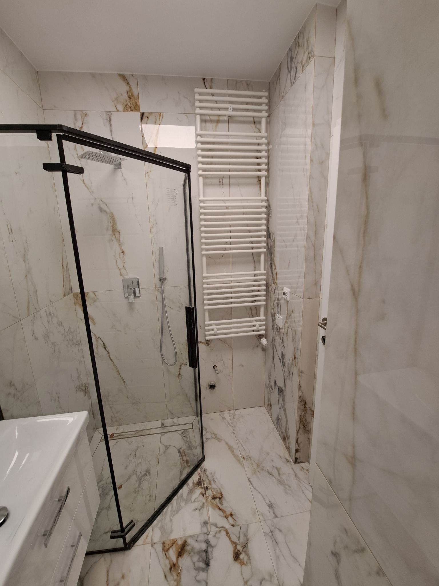 Nowoczesna łazienka z kabiną prysznicową typu walk-in z czarnymi profilami, marmurowe płytki na ścianach i podłodze, biały grzejnik drabinkowy.