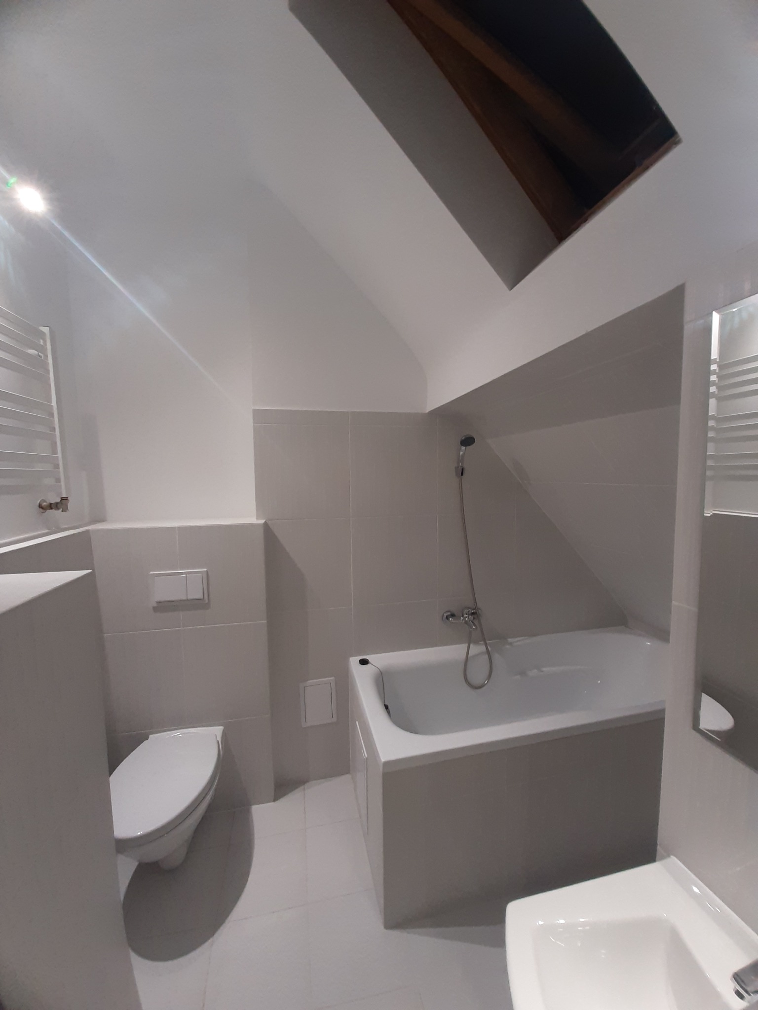 Jasna łazienka na poddaszu ze skośnym sufitem, biała wanna, umywalka i toaleta, grzejnik drabinkowy.