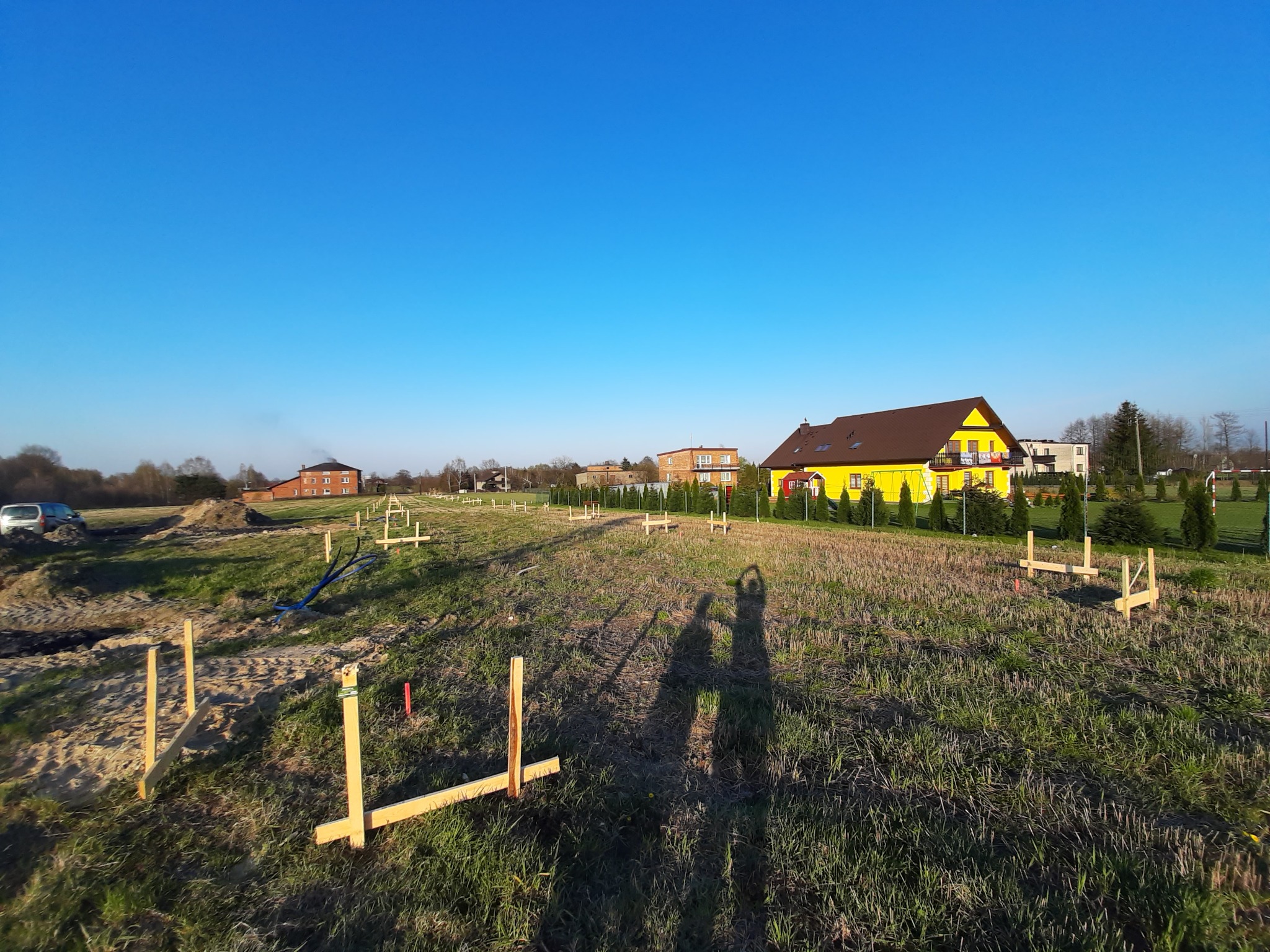 Wytyczanie fundamentów pod budowę domu jednorodzinnego; widoczne drewniane paliki i wstępnie przygotowany teren z zarysem przyszłego budynku, w tle żółty dom.