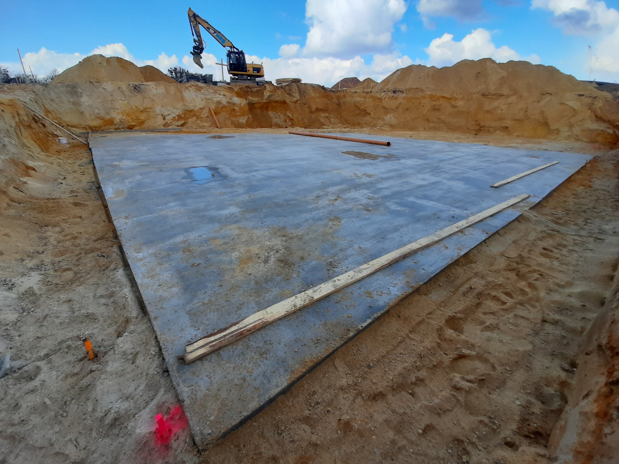 Wylana płyta fundamentowa na placu budowy z widocznym koparką na tle wykopanej ziemi i błękitnego nieba z chmurami.