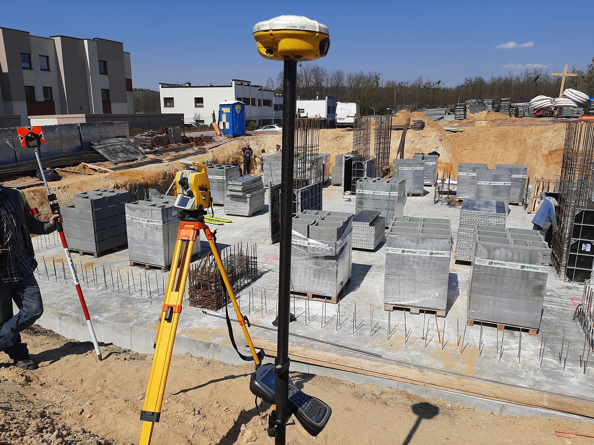 Budowa fundamentów budynku wielorodzinnego z użyciem niwelatora, tyczki i odbiornika GPS na statywie, widoczne bloki betonowe na paletach i zbrojenie.