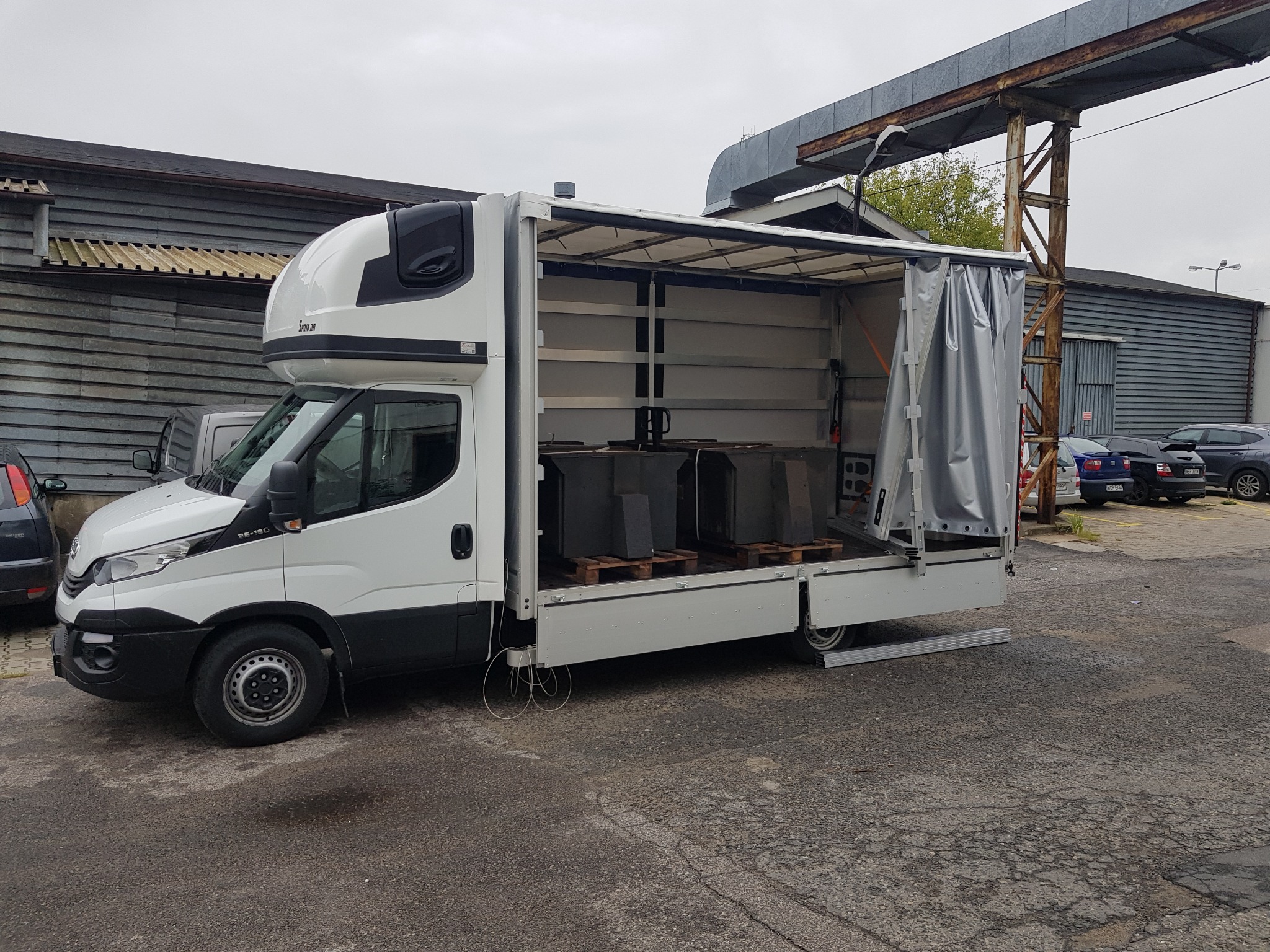 Biały samochód dostawczy z otwartą przestrzenią ładunkową, w której znajdują się cztery ciemne, prostopadłościenne ładunki na drewnianych paletach, stojący na szarym asfalcie na tle przemysłowych...