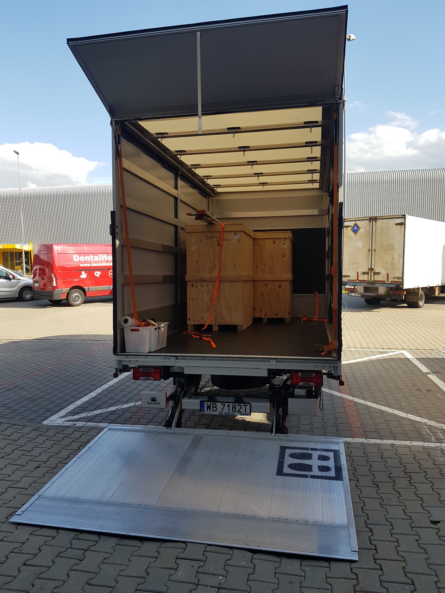 Otwarta paka busa z trzema drewnianymi skrzyniami zabezpieczonymi pomarańczowymi pasami, widoczna rampa załadunkowa i biały pojemnik z folią bąbelkową.