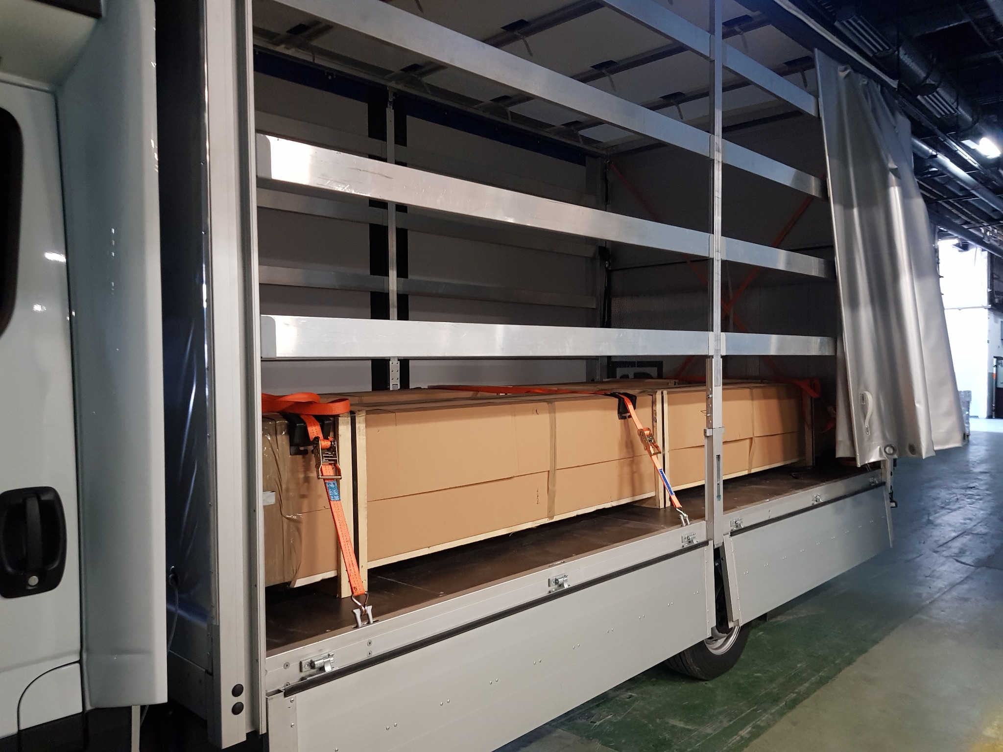 Wnętrze busa transportowego z ładunkiem kartonowych pudeł zabezpieczonych pomarańczowymi pasami transportowymi, widoczna metalowa konstrukcja wewnątrz przestrzeni ładunkowej.