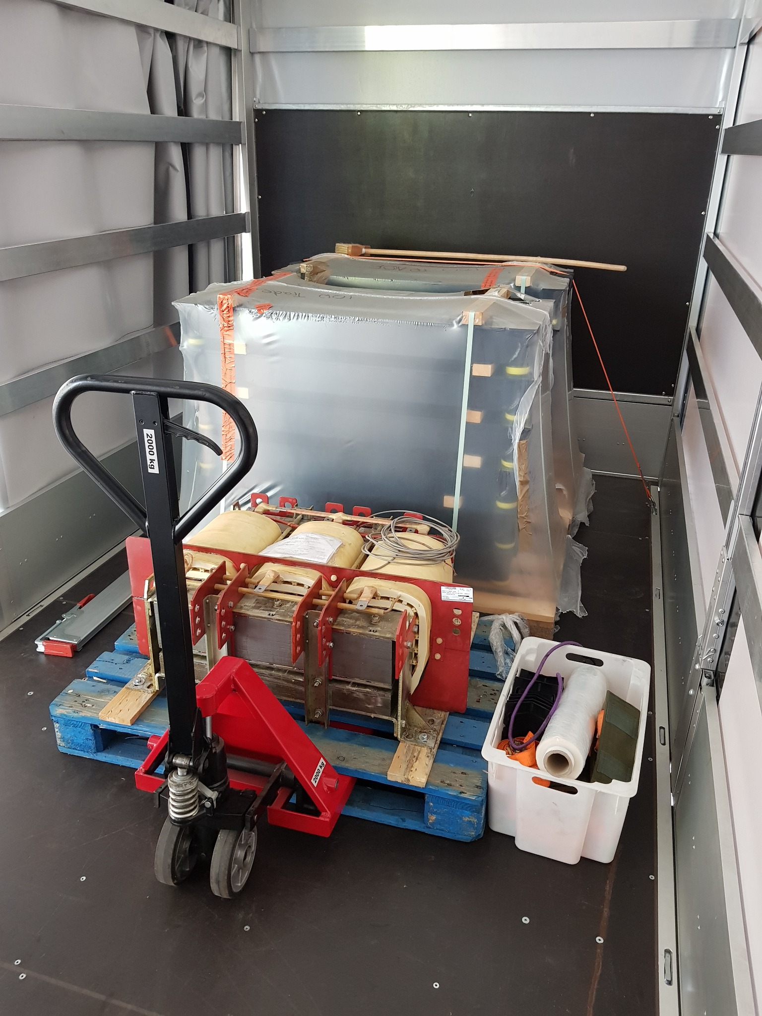 Transformator energetyczny zabezpieczony folią i pasami transportowymi, umieszczony na palecie w przestrzeni ładunkowej pojazdu, obok wózek paletowy i skrzynka z akcesoriami.