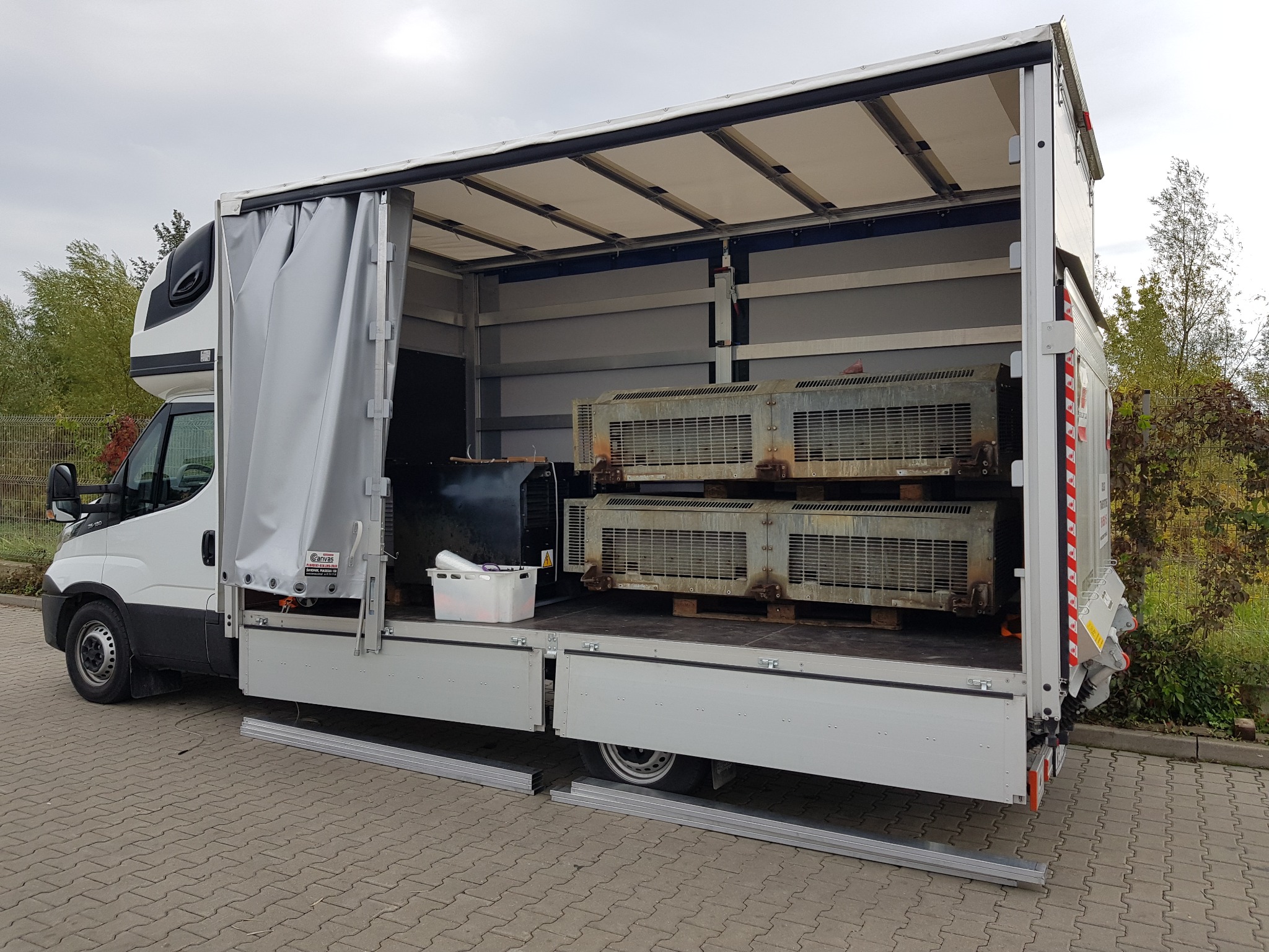 Otwarty biały samochód dostawczy z szarą plandeką, załadowany dwoma starymi, metalowymi klimatyzatorami na drewnianych paletach, widoczny biały pojemnik z zawiniętymi przedmiotami.