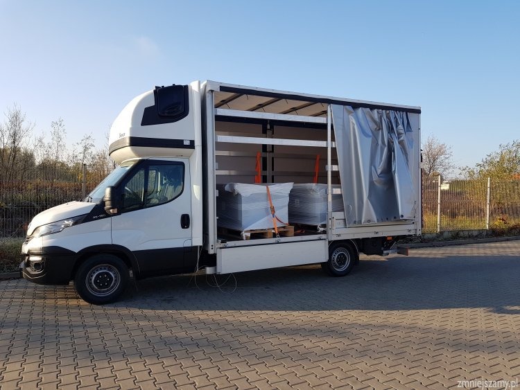 Biały samochód dostawczy typu bus z częściowo odsłoniętą plandeką, przewożący zapakowane palety zabezpieczone pasami.